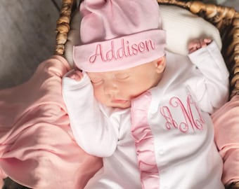 May include: A light pink knotted hat with the name Addison embroidered in pink thread, paired with a white long-sleeve onesie featuring a pink ruffled placket and a pink embroidered monogram on the chest, all resting on a soft pink fabric.