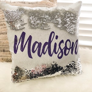 May include: A white pillow with silver sequins that can be flipped to reveal a black side. The pillow has the name "Madison" written in purple on the white side.