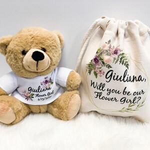 May include: A light brown teddy bear wearing a white t-shirt printed with purple floral graphics and the text Giuliana Flower Girl 6/13/2020, alongside a cream-colored drawstring canvas bag featuring a floral wreath and the text Giuliana, Will you be our Flower Girl?