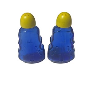 May include: Two blue glass salt and pepper shakers with yellow lids. The shakers are shaped like violins, with the strings and curves of the instrument visible. The yellow lids have small holes for dispensing the contents.