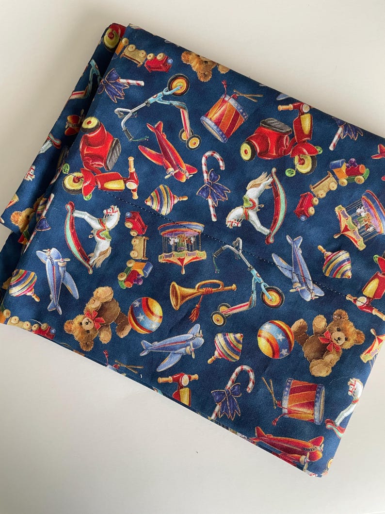 Op de afbeelding: Een blauwe stof met een herhalend patroon van vintage speelgoed, waaronder een schommelpaard, een speelgoedvliegtuig, een teddybeer, een speelgoedtrommel, een speelgoedtrein, een speelgoedstep en een speelgoedtol.