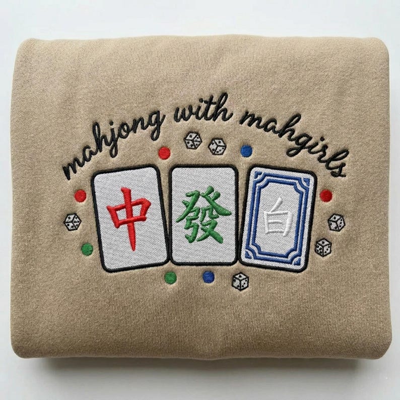 May include: Tan sweatshirt with embroidered Mahjong tiles and the text "mahjong with mahgirls" in black script. The tiles are white with red, green, and blue accents, and dice are scattered around the design.