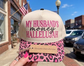 Puede incluir: Una gorra de béisbol color crema con una visera de estampado de leopardo rosa. El frente presenta el texto bordado MY HUSBAND'S money HALLELUJAH. Una etiqueta rectangular marrón unida a la visera muestra el texto ShopArchersArrow en negro.