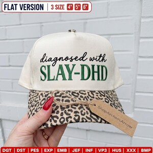 May include: A cream-colored baseball cap with a leopard print brim. The front features the embroidered black and green text 'diagnosed with SLAY-DHD'. A brown paper tag is attached to the brim, and the top text indicates 3 sizes: 4, 5, and 6 inches.