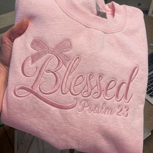 May include: A light pink sweatshirt with embroidered text and a bow design. The word "Blessed" is embroidered in a cursive font, with "Psalm 23" below. The sweatshirt is made of a soft material.