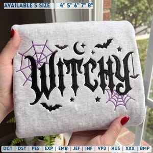Puede incluir: Una sudadera gris claro con el texto bordado en negro "WITCHY" en fuente gótica. El diseño incluye telarañas moradas, murciélagos negros, una luna creciente y estrellas. El texto superior indica los tamaños disponibles 4", 5", 6", 7", 8".