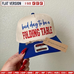 Op de afbeelding: Een crèmekleurige baseballpet met een blauwe klep, voorzien van de geborduurde tekst "bad day to be a FOLDING TABLE" in blauw en rood. De pet heeft een sticker op de klep en een bruin papieren label met "ShopArchersArrow" vastgebonden met touw.