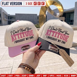 May include: Two baseball caps with cream-coloured crowns and embroidered text reading I Don't Need Your ATTITUDE I BROUGHT MY OWN. One cap has a pink brim, the other a black brim. A tag labelled ShopArchersArrow is attached to the black-brimmed cap.