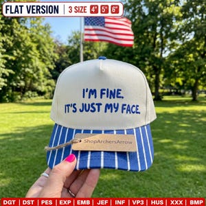 May include: A cream-coloured baseball cap with blue embroidery that reads "I'M FINE. IT'S JUST MY FACE." The brim features a blue and white vertical stripe pattern. A brown rectangular tag with the text "ShopArchersArrow" is attached to the brim.