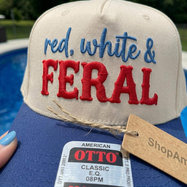 K&ouml;nnte beinhalten: Eine beige-blaue Baseballkappe mit dem gestickten Text red, white & FERAL in blauem und rotem Garn. Die Kappe enth&auml;lt einen rechteckigen Aufkleber mit dem Text AMERICAN OTTO CLASSIC E.Q. 08PM und ein braunes Papieretikett, das mit einer Schnur befestigt ist.