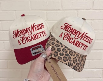 Puede incluir: Dos gorras de béisbol con coronas color hueso y texto bordado en rojo Mommy Needs A Cigarette. Una gorra tiene una visera roja sólida, mientras que la otra presenta una visera con estampado de leopardo marrón y negro. Texto 10cm 13cm 15cm visible.