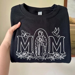 May include: Black sweatshirt with the word "MOM" in white script, featuring a religious illustration in the center. A butterfly and a dove are on either side of the word. Roses are at the bottom of the letters.