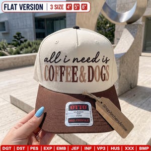 May include: A cream-colored baseball cap with a brown brim, featuring the embroidered text "all i need is COFFEE & DOGS" in black and brown. A rectangular sticker on the brim reads "OTTO CLASSIC FIT OSFM," and a brown tag reads "ShopArchersArrow."