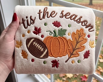 May include: A cream-colored fabric featuring an embroidered design with the text 'tis the season' in brown. The graphic includes a brown football, an orange pumpkin, and various red, orange, and yellow autumn leaves, available in 4, 5, 6, 7, and 8 inch sizes.