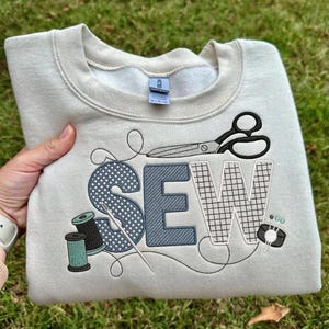 Pode incluir: Um moletom bege claro com a palavra "SEW" bordada em um design temático de costura. O design inclui tesouras, carretéis de linha e uma agulha. O moletom é exibido na grama verde.