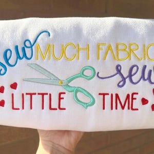 May include: White fabric with the embroidered phrase "Sew Much Fabric, Sew Little Time" in blue, yellow, and purple. A pair of teal scissors and red hearts are also embroidered on the fabric.