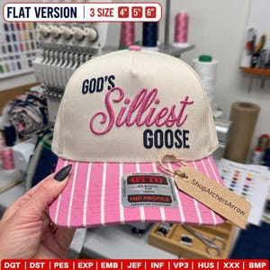 May include: A cream-colored baseball cap with a pink and white striped brim. The front features embroidered text reading "GOD'S" in navy, "Silliest" in pink script, and "GOOSE" in navy. A brown paper tag is attached to the brim with twine.
