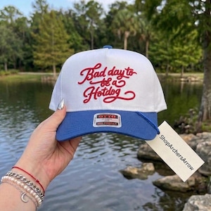 May include: A white and blue baseball cap with the text "Bad day to be a Hotdog" in red script. The cap has a blue brim and a blue button on top. The cap is held up by a hand.