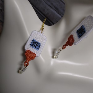 May include: Embroidered glue bottle earrings with gold-colored hooks and clear bead accents. The white earrings feature the word "GLUE" in blue, with orange and brown details. The earrings are displayed on a mannequin.
