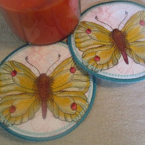 May include: Two round coasters with a teal border and a white background. Each coaster features an embroidered yellow butterfly with red accents.
