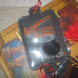 May include: A gray leather bookmark with the text "Advanced Potion-Making" in orange embroidery. The bookmark has a clear plastic cover and a red tassel.