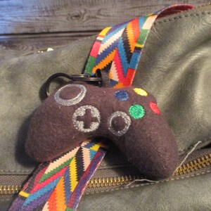 May include: A dark grey plush toy shaped like a video game controller with colourful buttons. The toy is attached to a colourful striped strap.