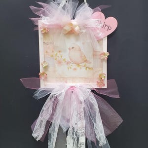 May include: A pink and white bird-themed door hanger with a pink bow, lace, and tulle. The hanger features a pink bird on a branch with pink flowers. The word "Chirp" is written on a pink heart.