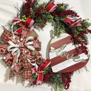 May include: A red and white striped heart-shaped wreath with a metal sign that says "love" and a bow made of red, white, and brown ribbon. The wreath is decorated with red berries and green leaves.