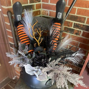May include: A Halloween decoration featuring a black cauldron filled with decorative elements. The cauldron holds witch legs with orange and black striped stockings, black boots, and various faux foliage in shades of black, gray, and gold. A festive holiday display.