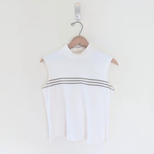 May include: A white sleeveless top with a high neck and two grey stripes across the chest. The top is displayed on a wooden hanger. The background is a plain white wall.
