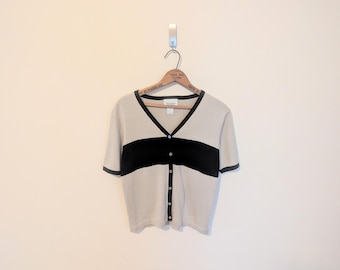 May include: A beige short-sleeved cardigan with a black horizontal stripe across the chest. The cardigan has a V-neck, black trim, and a button-up front. It hangs on a wooden hanger against a plain wall.