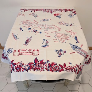 May include: A rectangular off-white textile featuring a red and blue printed map of Hawaii. The design includes illustrations of surfers, hula dancers, and tropical flowers, with text reading Map of Hawaii, Pacific Ocean, and Aloha across the surface.