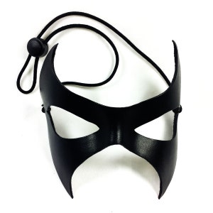 K&ouml;nnte beinhalten: Eine schwarze Leder-Maskenballmaske mit spitzem Design und Augenausschnitten. Die Maske ist an einem schwarzen Gummiband befestigt.