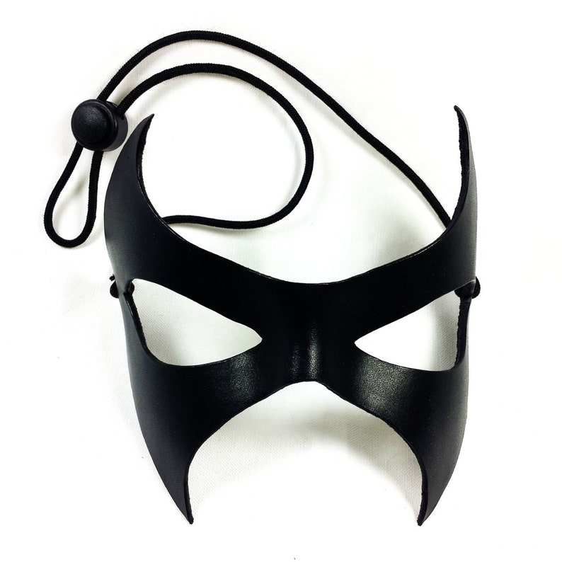 K&ouml;nnte beinhalten: Eine schwarze Leder-Maskenballmaske mit spitzem Design und Augenausschnitten. Die Maske ist an einem schwarzen Gummiband befestigt.