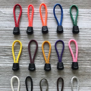 May include: A collection of colorful zipper pulls in various shades, including red, orange, blue, green, yellow, purple, and pink. Each pull features a loop of cord attached to a black plastic base. The pulls are arranged on a light wood-grain surface.