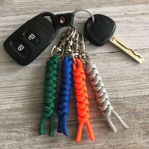 May include: A collection of keys and keychains. The image features a black car key fob, a car key, and a set of four braided paracord keychains in green, blue, orange, and gray. The keychains are attached to a metal ring.