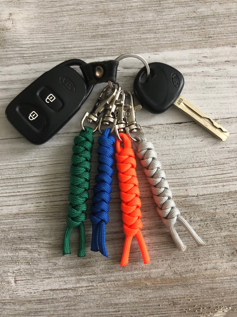 May include: A collection of keys and keychains. The image features a black car key fob, a car key, and a set of four braided paracord keychains in green, blue, orange, and gray. The keychains are attached to a metal ring.