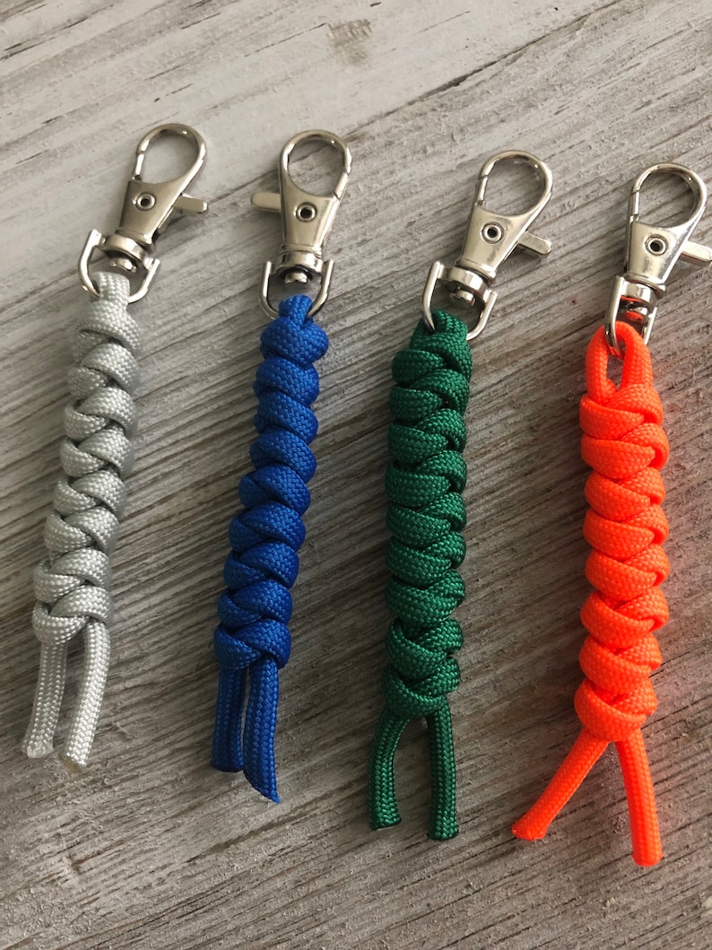 May include: Four paracord keychains in silver, blue, green, and orange. Each keychain features a woven cord design and a silver-colored metal clasp. The cords are tightly woven, with the ends of the cord extending below the woven section.