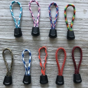 May include: A collection of ten zipper pulls in various colors and patterns. The pulls feature a black plastic base with a loop made of braided cord. Colors include blue, pink, red, green, and orange. The cords have patterns like camouflage and geometric designs.