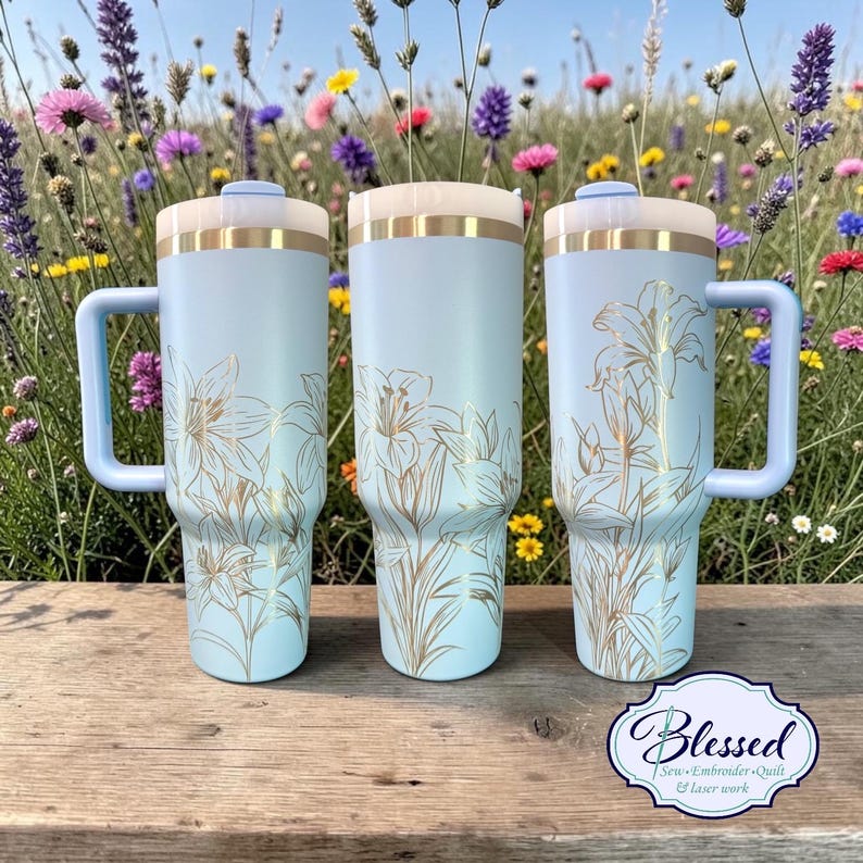 May include: Three light blue insulated tumblers with handles, featuring a gold floral design. Each tumbler has a light blue handle, a white top, and a gold band. The tumblers are displayed on a wooden surface with a colorful flower background.