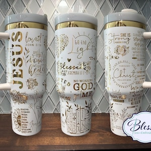 May include: Three white insulated tumblers with gold accents and handles. Each tumbler features gold script and floral designs with religious quotes, including "Jesus," "Be the Light," and "Strong."
