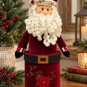 May include: A cylindrical Santa Claus figurine featuring a deep red velvet body and hat. The figure has a thick, curly white yarn beard, a black belt with a gold buckle, and a base decorated with embroidered red poinsettia flowers and vines.