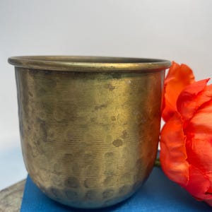 May include: A vintage brass planter with a hammered texture. The cylindrical vessel has a rounded base and a slightly flared rim. An orange flower rests beside the planter on a blue surface.