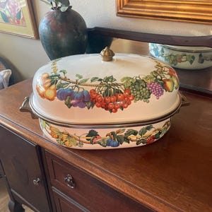 May include: An oval-shaped white enameled roasting pan with a matching lid, featuring a decorative fruit garland pattern of pears, plums, cherries, and grapes. The pan includes brass-toned metal side handles and a small brass-toned knob on the lid.