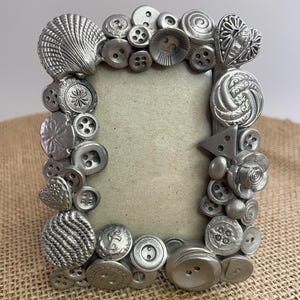 May include: A rectangular picture frame featuring a border decorated with various silver-toned buttons and decorative shapes, including a large seashell, a heart, and a dollar sign, surrounding a plain, light-colored rectangular center insert.