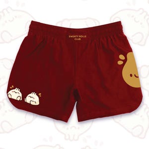 Peut inclure: Short de sport bordeaux avec le texte "SWEATY ROLLS CLUB" et des graphiques de raviolis. Le short a une taille élastique et une coupe décontractée. Un grand graphique de ravioli est sur la jambe droite et deux plus petits sur la jambe gauche.