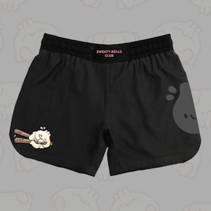 Könnte beinhalten: Schwarze Sportshorts mit elastischem Bund und dem in Pink aufgedruckten Text SWEATY ROLLS CLUB. Auf dem linken Bein befindet sich eine kleine Grafik eines Cartoon-Teigtaschen-Wesens mit Stäbchen, rechts eine große, blasse graue Klecksform.