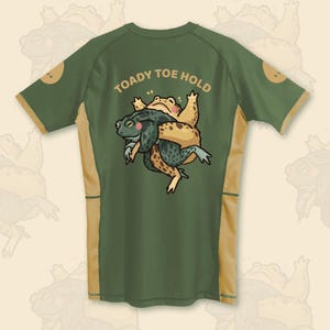 Op de afbeelding: Een olijfgroen shirt met korte mouwen en beige zijpanelen. De achterkant toont een afbeelding van een donkergroene pad die een lichtbeige pad vasthoudt, met de tekst TOADY TOE HOLD in een beige boog boven de twee afgebeelde kikkers.