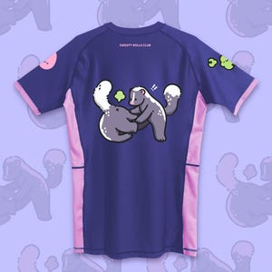 May include: A navy blue athletic shirt with pink accents and a cartoon skunk graphic. The shirt features the text "SWEATY ROLLS CLUB" on the back. The sleeves and sides have pink trim, and a pink circle and green shapes are on the sleeves.