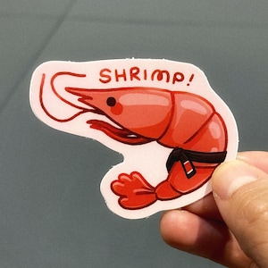 May include: A sticker depicting a cartoon shrimp in red, wearing a black belt, with the word "SHRIMP!" in red. The shrimp has a smiling face and is on a white background.